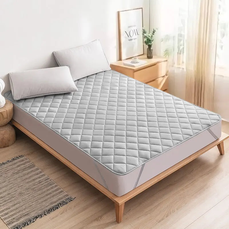 Cubrecolchón acolchado gris claro en cama moderna con almohadas, dormitorio luminoso y decoración minimalista.