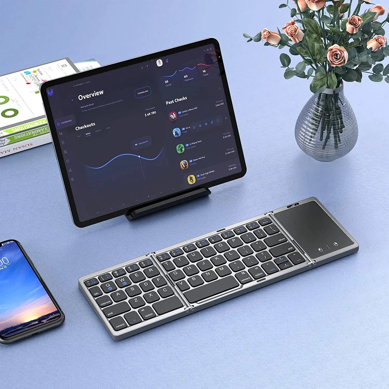 Teclado plegable inalámbrico con touchpad junto a tablet, smartphone y jarrón de flores en escritorio moderno.